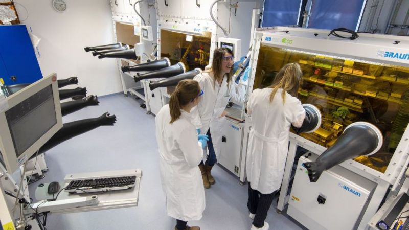 Drei Wissenschaftlerinnen in weißen Kitteln und mit Schutzbrille stehen in einem länglichen Gang mit verschiedenen technischen Laborapparaturen.