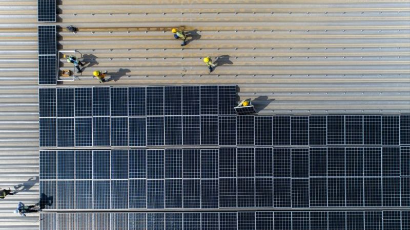Symbolfoto: Arbeiter bei der Installation von Photovoltaikmodulen auf einem Fabrikdach aus der Vogelperspektive.
