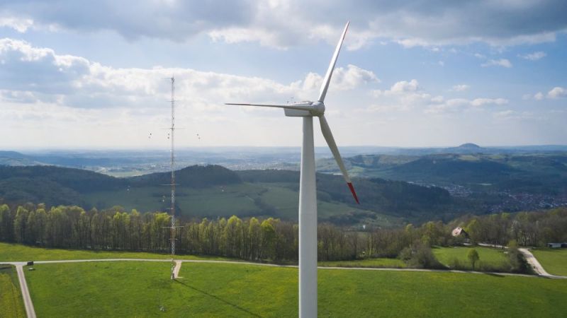 Eine Windenergieanlage im Vordergrund, im Hintergrund sieht man einen Windmessmast und eine hügelige Landschaft.