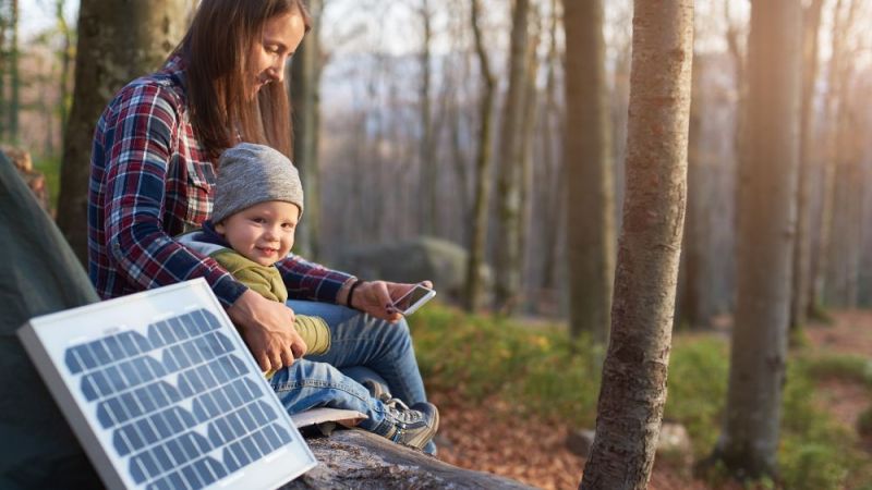 Symbolfoto: Ein kleiner Junge sitzt zusammen mit seiner Mutter in einem herbstlichen Wald auf einer Bank, auf der ein mobiles Solarmodul steht.