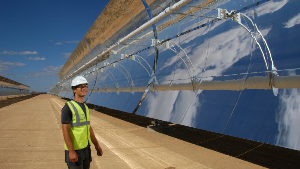 Ein Mann steht vor großen Parabolrinnenspiegeln eines Kraftwerke. Diese  konzentrieren die Sonnenstrahlen auf eine Rinne, in der ein flüssiges Wärmeträgermedium zirkuliert.