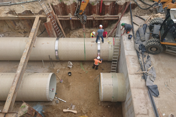 Auf dem Foto ist eine Baugrube zu sehen, in der ein Rohr liegt, auf dem ein Arbeiter steht.